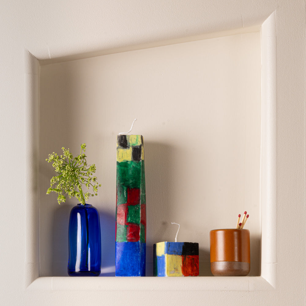 A built in shelf in a wall, with hand painted rectangular tall and short candles in a colorful checkerboard pattern, a blue vase with a stem of greenery, and a small ceramic vessel with matches.