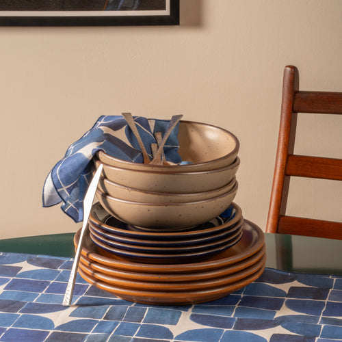 Stacked ceramic bowls and plates with blue patterned napkins and silver flatware on a dark green table.