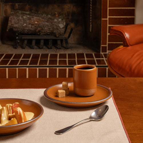 A rustic earthy orange ceramic cup filled with coffee sits on a matching saucer with two sugar cubes, set on a wooden table in a cozy room with a brick fireplace and an orange leather chair in the background.