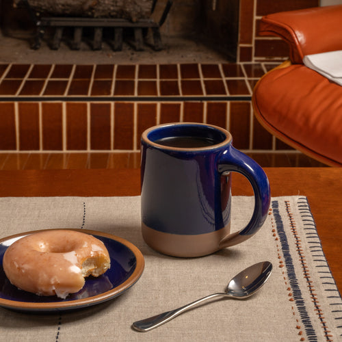 A large dark glossy blue ceramic mug filled with coffee sits on a wooden table next to a glazed donut on a matching blue plate, with a spoon on a placemat in front of a fireplace.
