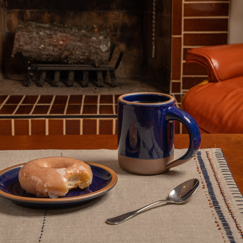 Glossy dark blue ceramic mug and plate with a glazed doughnut and spoon on a linen tablecloth in front of a fireplace.