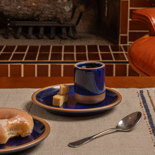A tiny ceramic cup and matching saucer in a dark glossy blue glaze with a natural beige clay edge, styled on a woven placemat in a cozy kitchen setting with a spoon and a pastry.