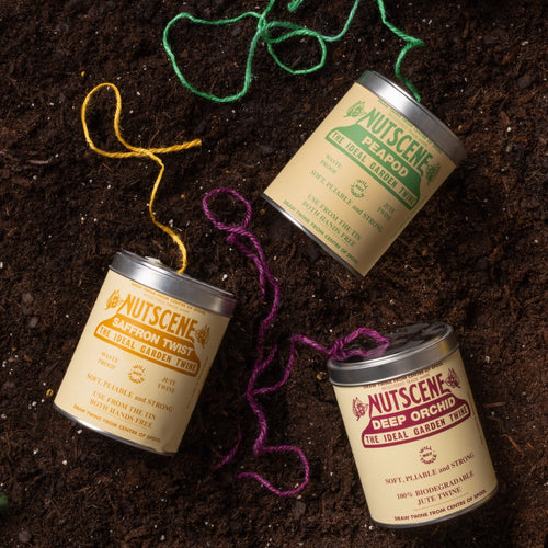 Three tins of garden twine (in green, yellow, and purple) placed on soil, with strands of colored twine loosely unspooled around them.