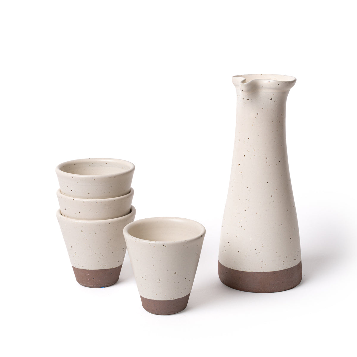 A tall ceramic carafe and stack of 4 tapered cups in an off white color, featuring iron speckles and unglazed bases.