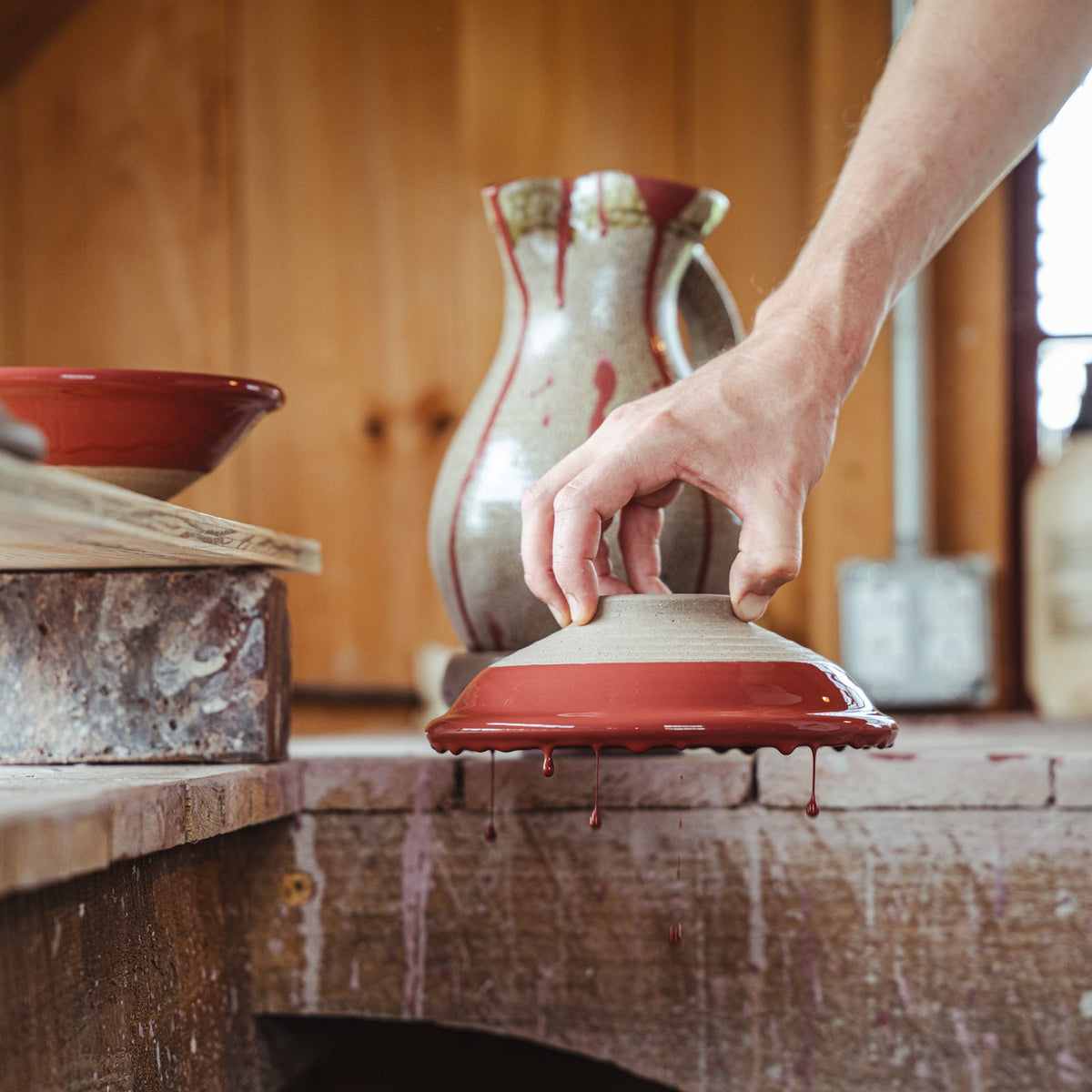 In a pottery studio, a hand holds a bowl upside down for glazing, as the glaze drips off.