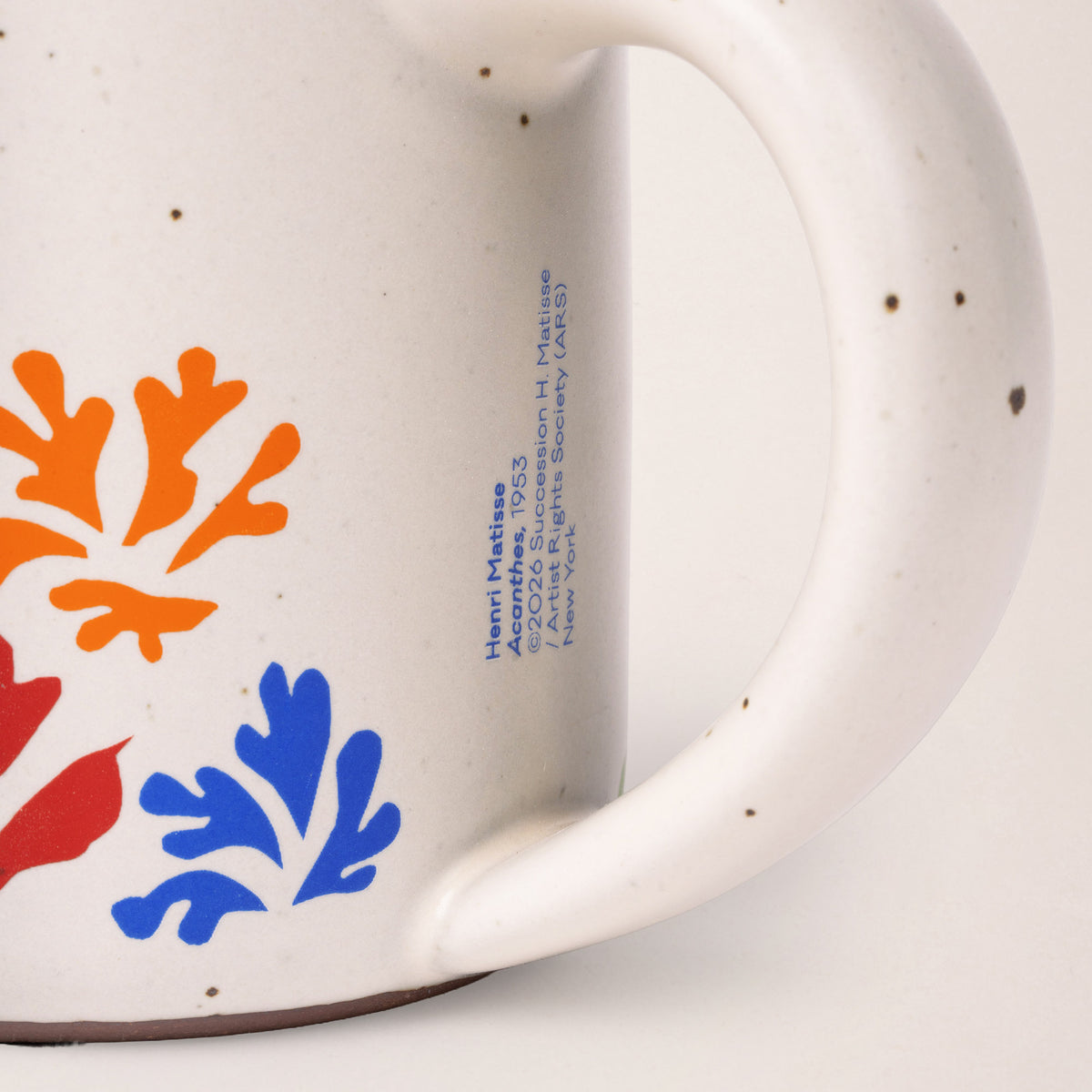 Close-up of a speckled off-white ceramic mug showing part of the body, a handle, blue printed text, and orange and blue floral motifs on a white background.