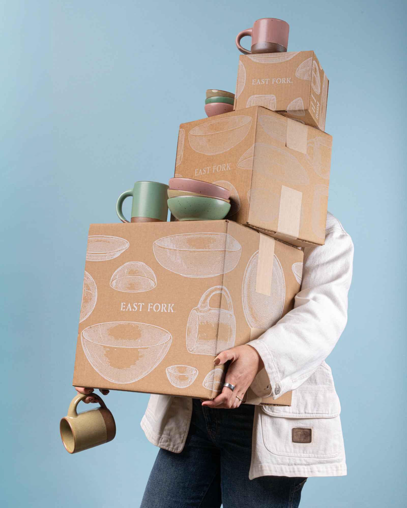 Person holding a stack of 'East Fork' branded boxes with mugs and bowls on a blue background