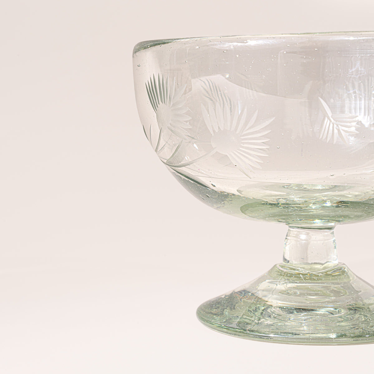 A closeup of a clear etched coupe glass with a pedestal base and delicate botanical pattern, shown on a clean neutral background.
