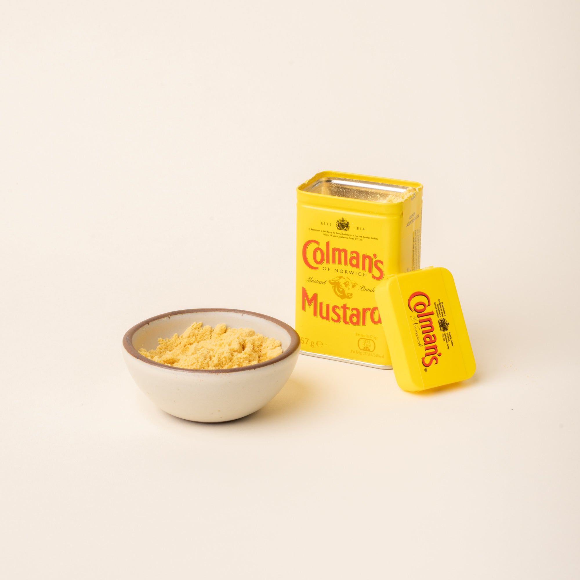 Open tin of Colman's Mustard Powder beside a ceramic bowl filled with mustard powder on a plain white background.