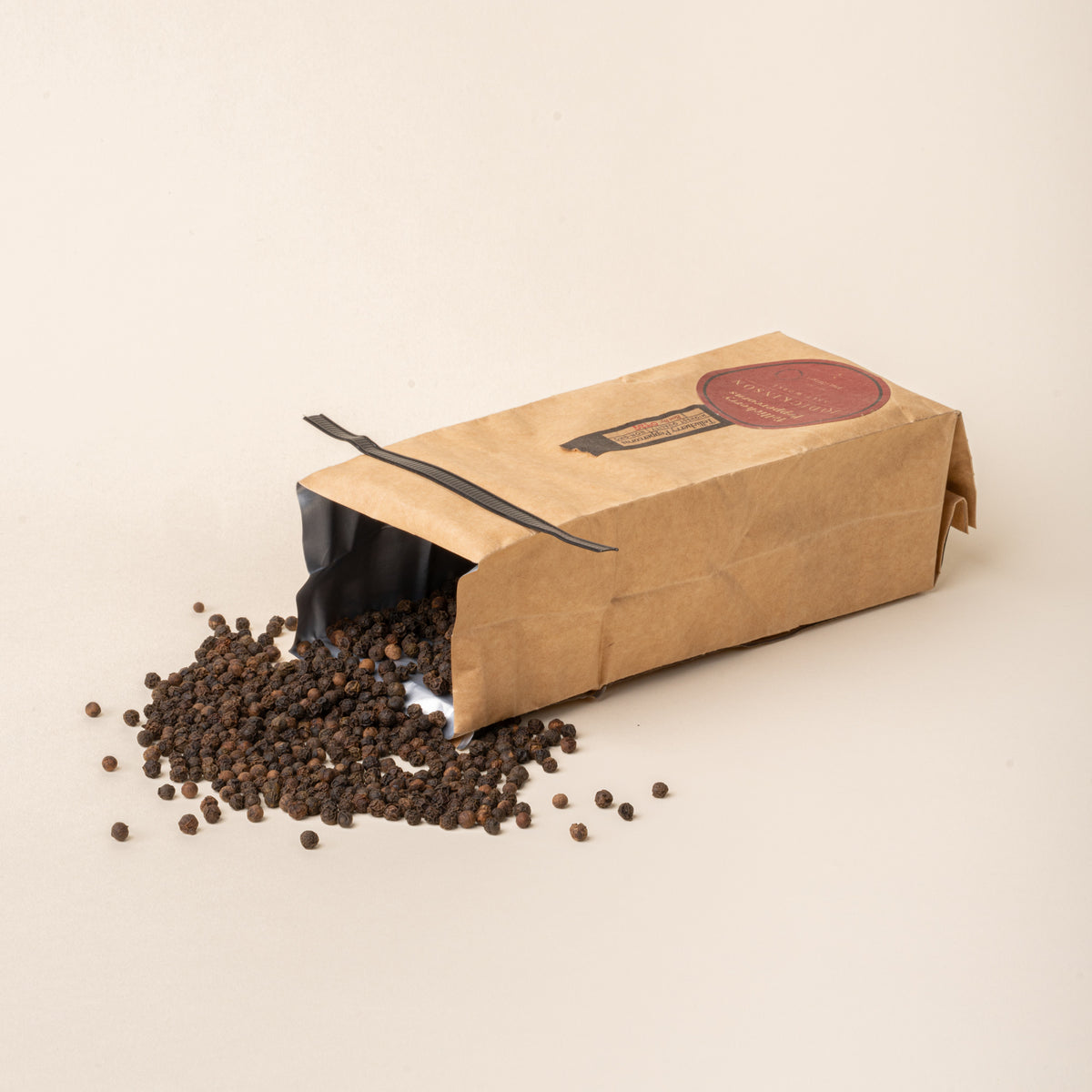 Opened bag of J.Q. Dickinson Tellicherry Peppercorns with whole black peppercorns spilled onto a plain white background.