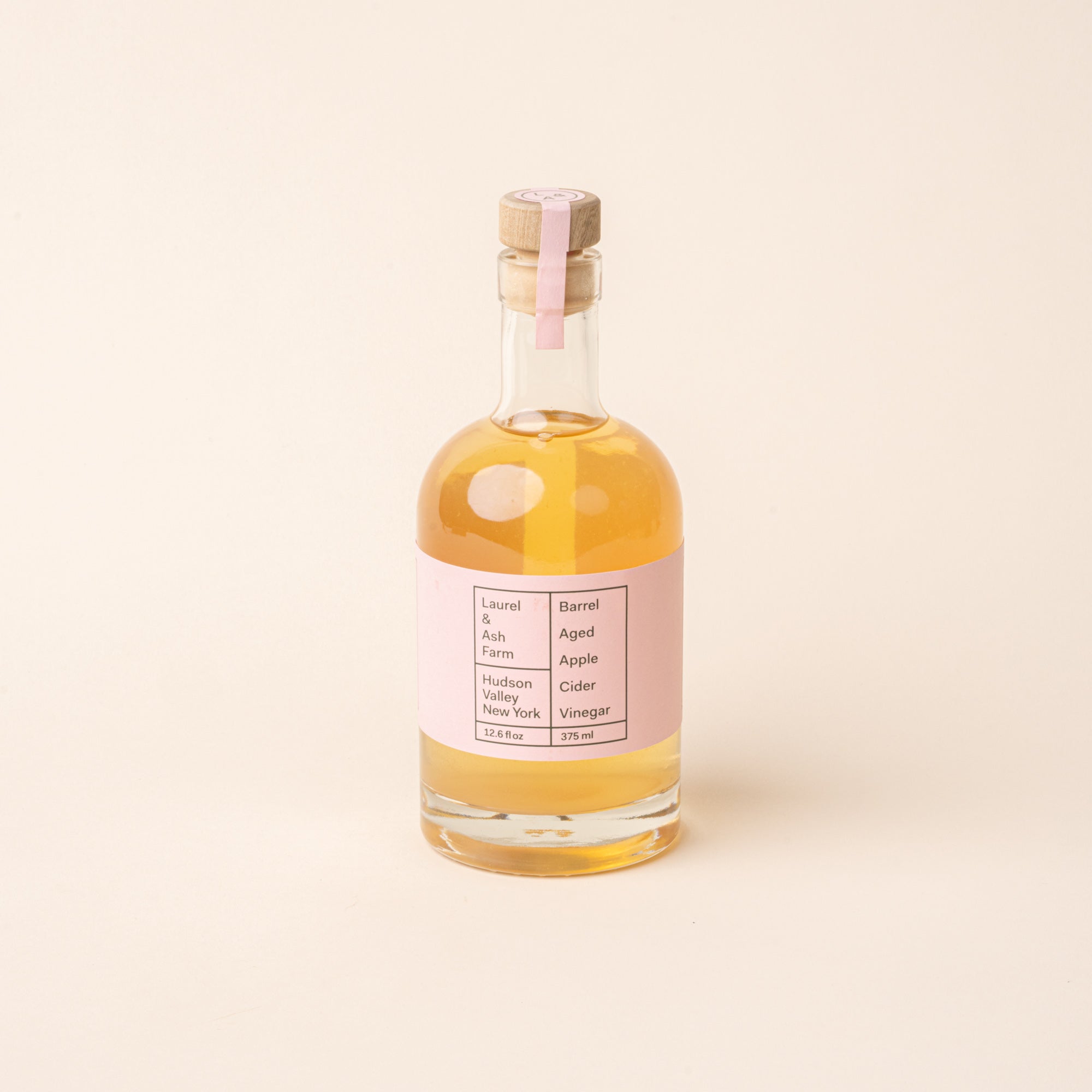 Bottle of Laurel & Ash Farm barrel-aged apple cider vinegar with a pink label and wooden stopper on a plain white background.