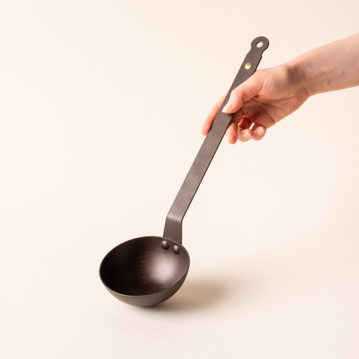 A hand holds out a black metal ladle with cup and a hole at the top of the handle