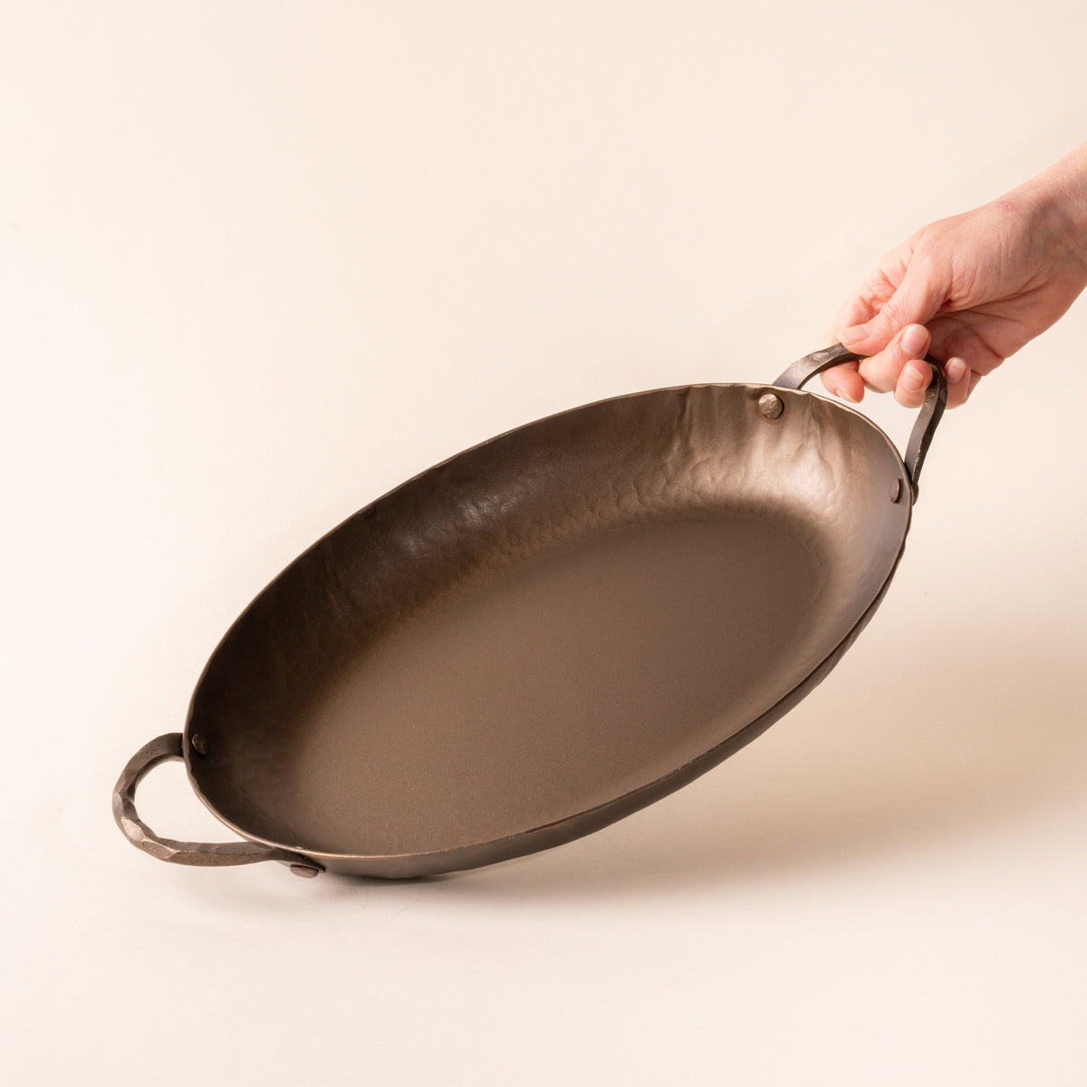 A hand tilting a hammered metal oval roasting pan with handles on both sides