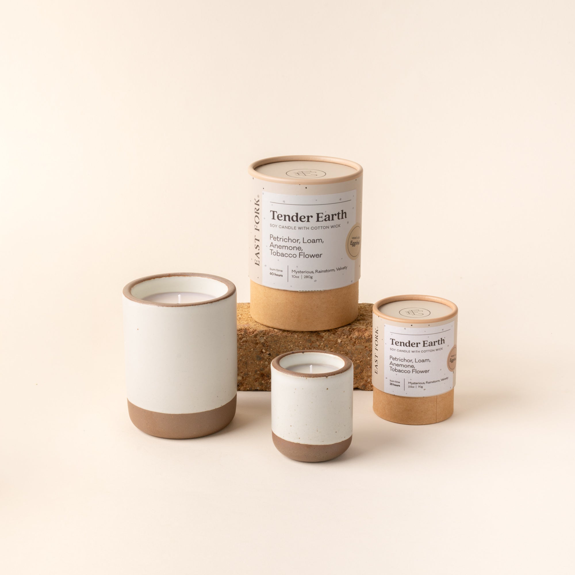 Small and large ceramic vessel in a cool white color with candles inside, arranged with tube packaging that reads 'Tender Earth'