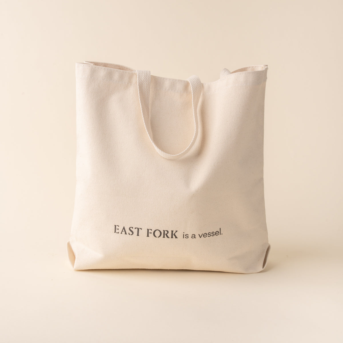 Back of a beige cotton tote bag with a black printed that reads 'EAST FORK is a vessel.'.