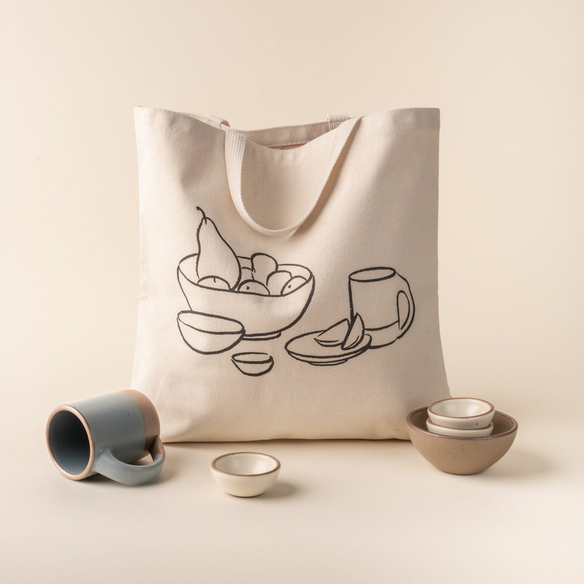 Tote bag with a drawing of a bowl of fruit and cups, surrounded by ceramic items on a beige background
