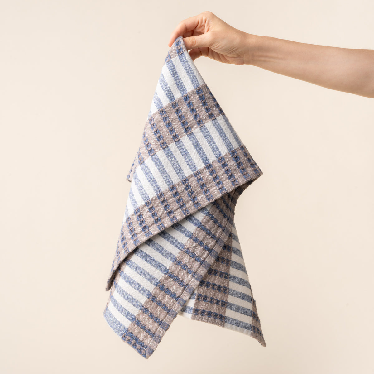 Hand holding out a woven kitchen towel with blue and beige striped pattern on a light background.