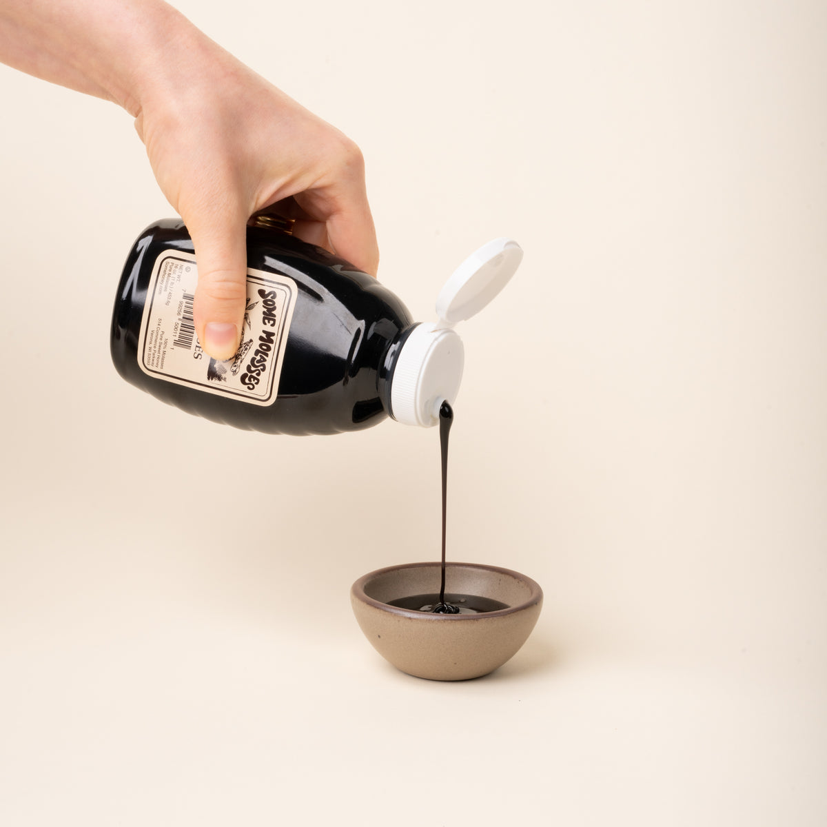 Hand pouring pure molasses from a bottle into a small ceramic bowl.