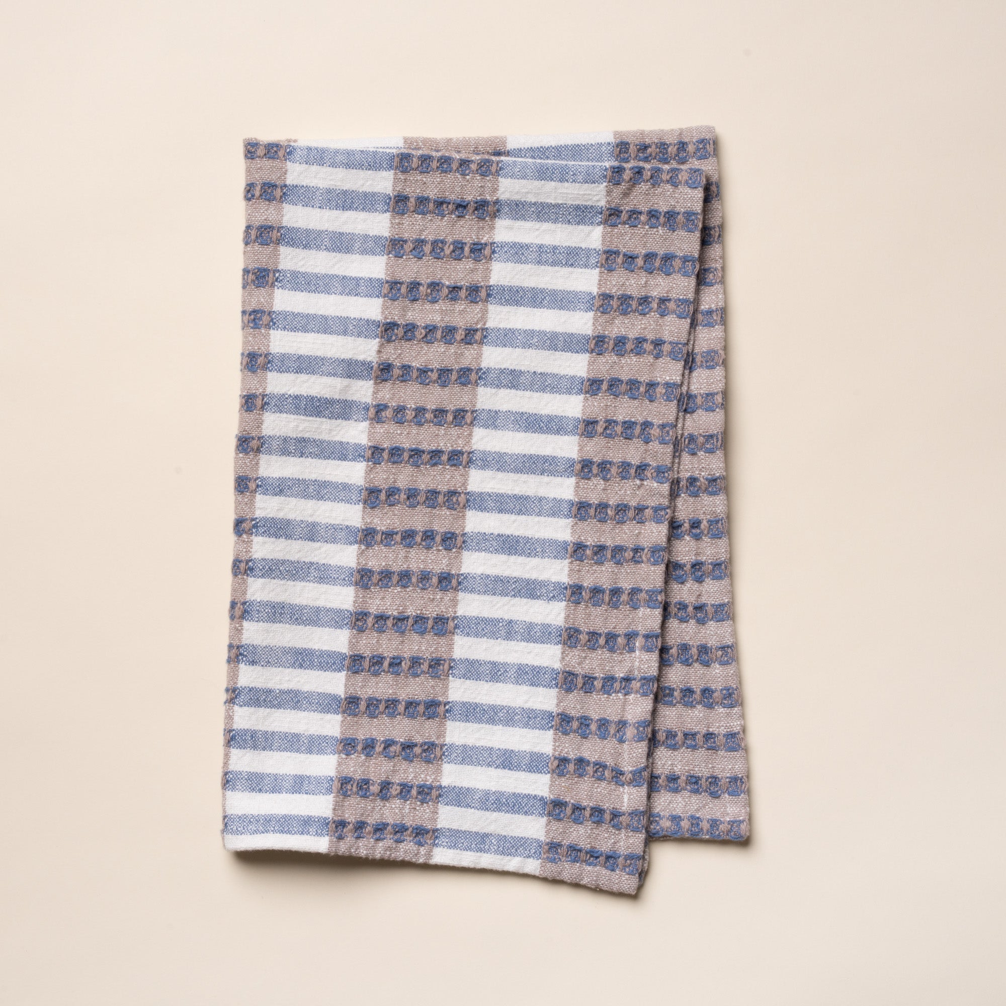 Folded woven kitchen towel with blue and beige striped pattern on a light background.