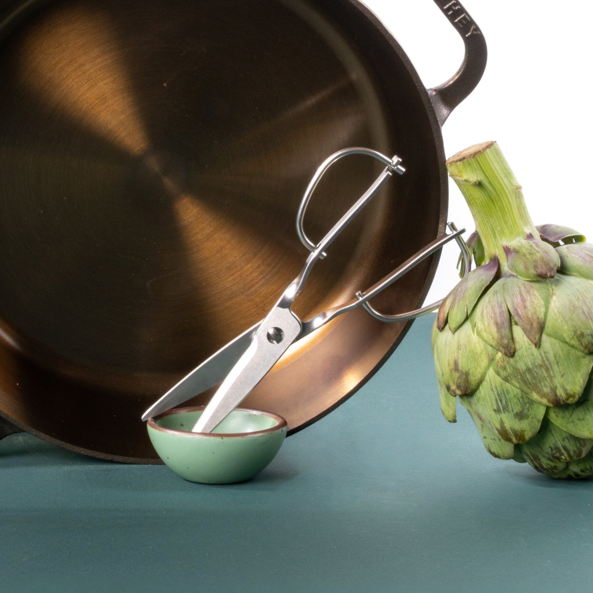Close-up of a skillet pan, kitchen scissors, and an artichoke beside a small green bowl