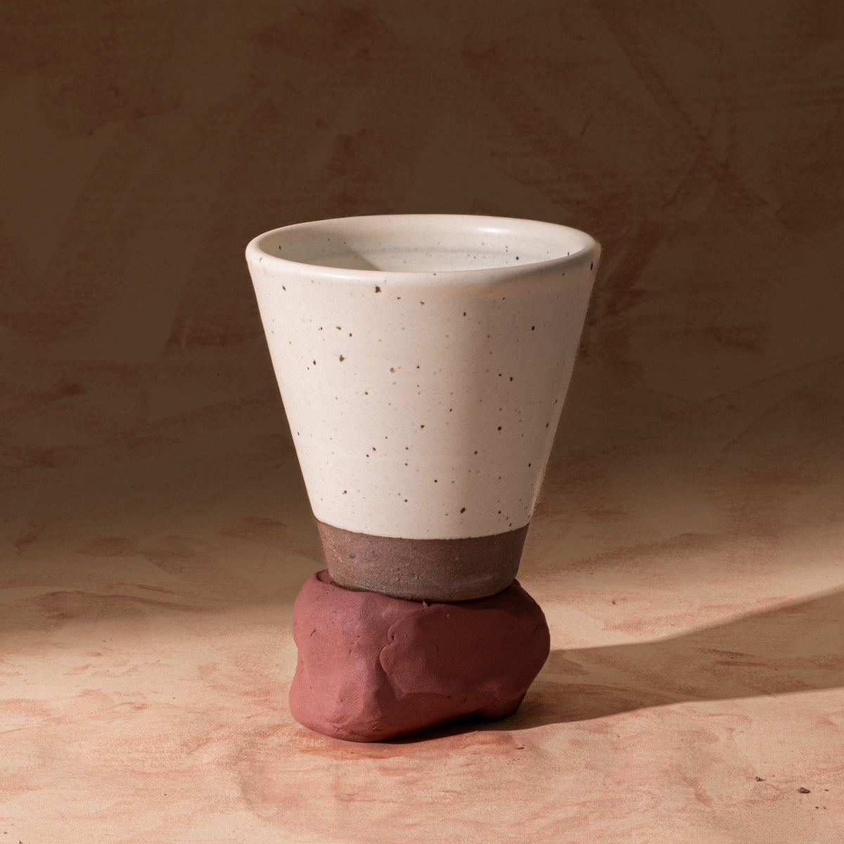 A tapered cup in an off-white color with iron speckles and unglazed base, sitting on raw clay.