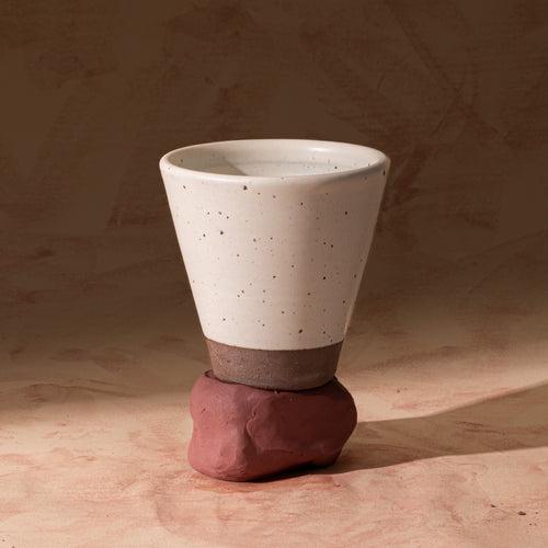 A tapered cup in an off-white color with iron speckles and unglazed base, sitting on raw clay.