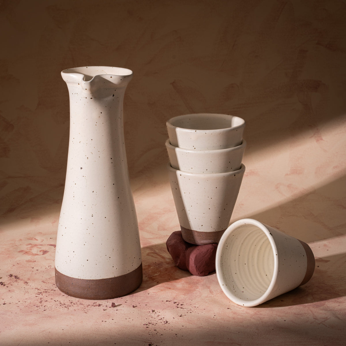 A tall ceramic carafe and stack of 4 tapered cups in an off white color, featuring iron speckles and unglazed bases, with some raw clay.
