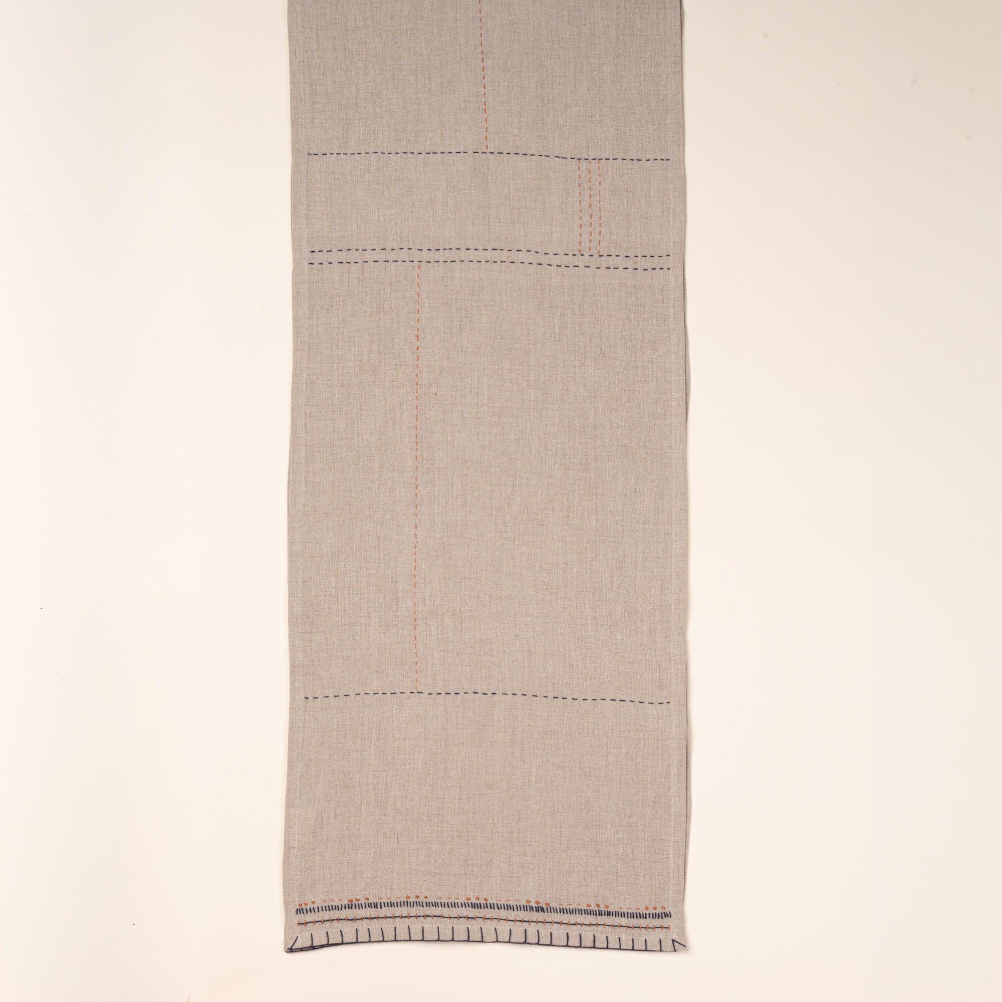 A neutral table runner, partially folded to showcase contrasting hand-stitched details in navy and rust.