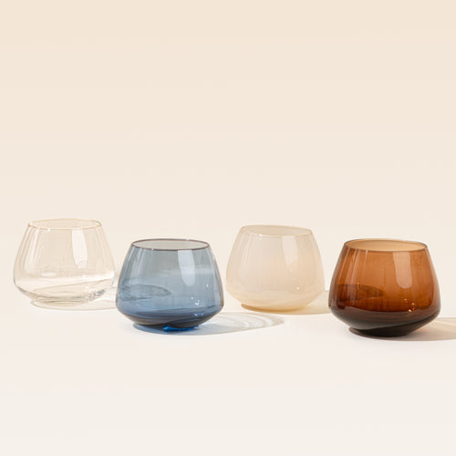 Four small stemless glass tumblers in different colors are lined up on a light beige background: clear, smoky blue, frosted cream, and smoky amber brown.