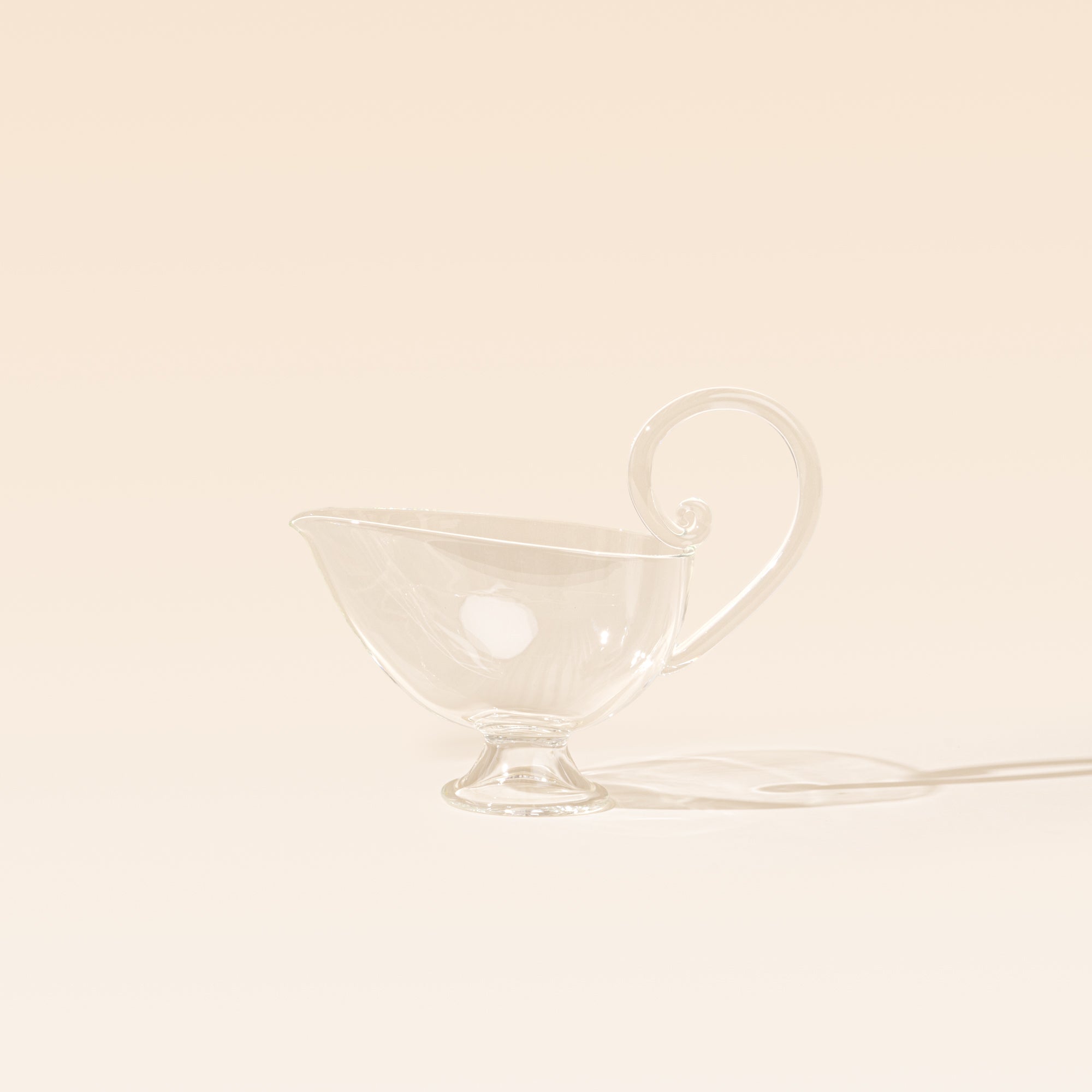 A clear glass sauce boat with a delicate curled handle, pointed pouring spout, and small pedestal foot, photographed on a warm cream background.