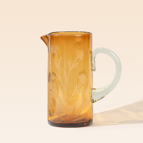 A large glass amber pitcher with handle made of recycled glass with etched thistle designs on it.