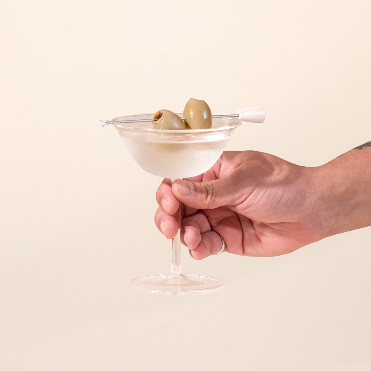 A hand holds out a clear martini coupe glass, filled with a martini with olives on a cocktail pick.
