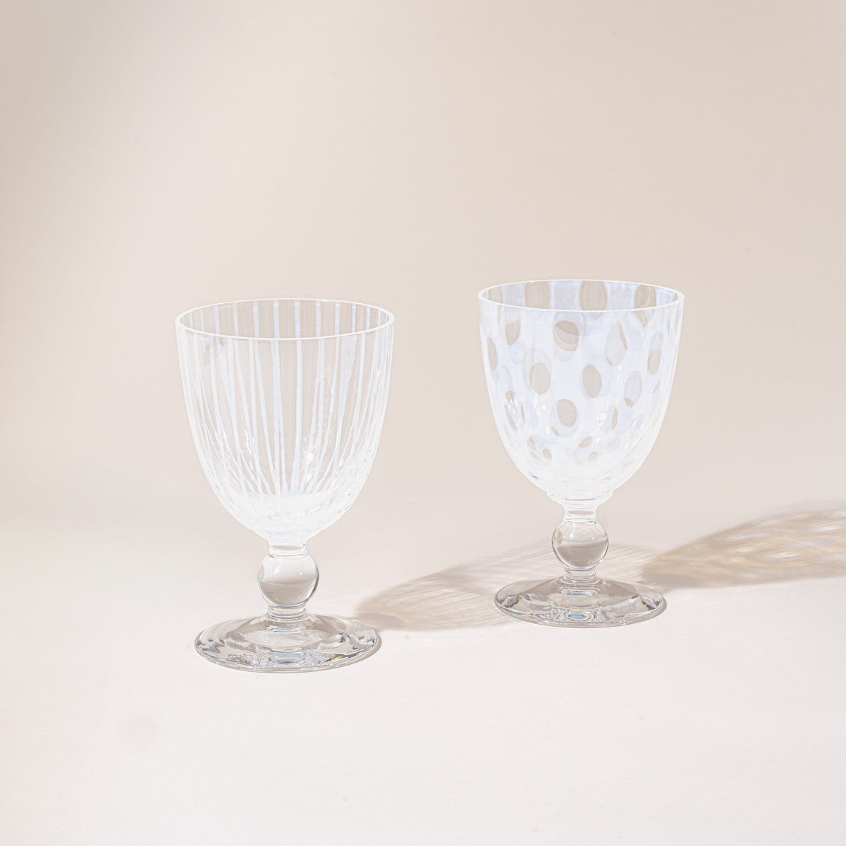 Two clear stemmed goblets shown side by side, one with vertical ribbing and one with a polka-dot pattern, photographed against a warm neutral background.