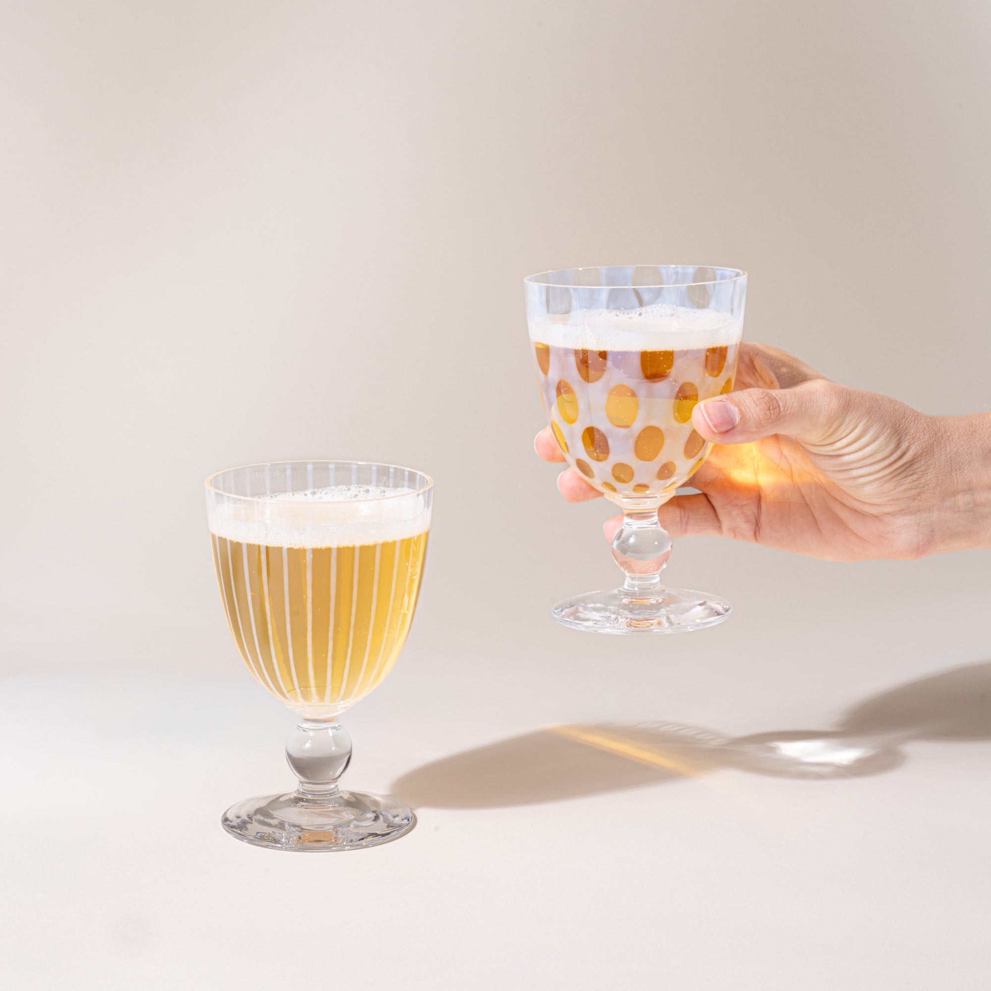Two clear patterned stemmed goblets filled with a pale golden sparkling drink, one resting on the table and one held in a hand, against a warm neutral background.