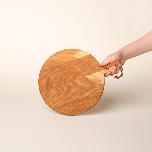 A hand holds a small round cherry serving board with a short handle and leather hanging loop, shown held against a warm cream background.