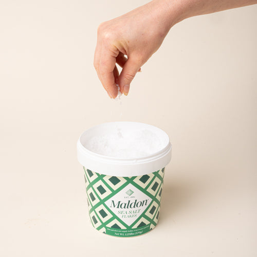 A hand sprinkling Maldon Sea Salt Flakes into an open green-and-white patterned container.
