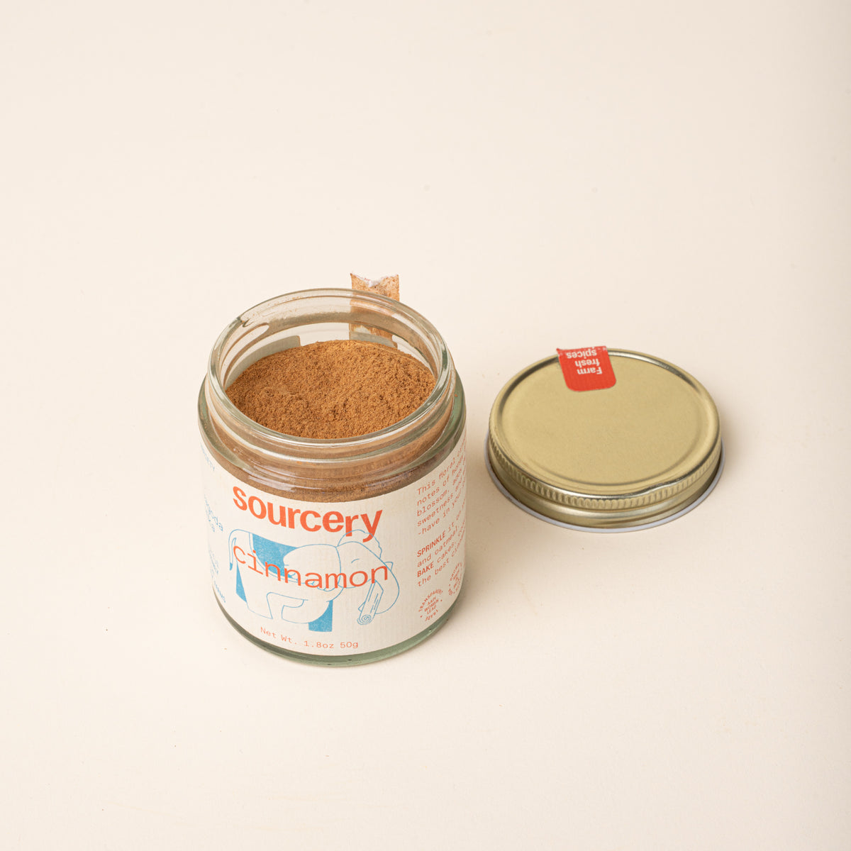 Glass jar of Sourcery cinnamon with a cream label featuring orange lettering and a blue elephant illustration, open with a old metal lid to the side.