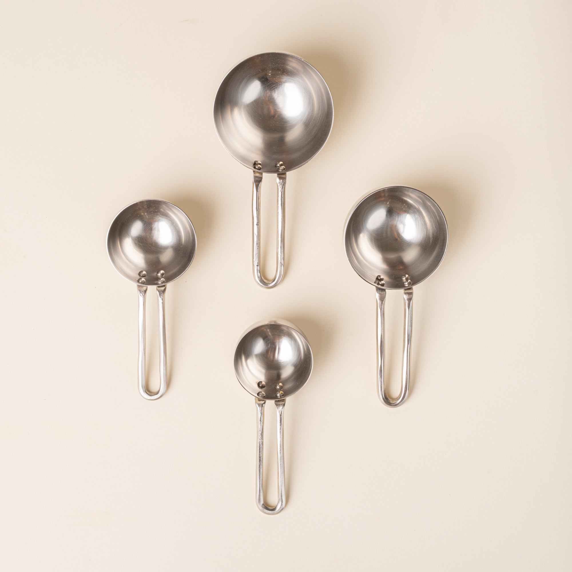 Four forged measuring cups in graduated sizes, arranged in a loose diamond formation on a warm cream background.
