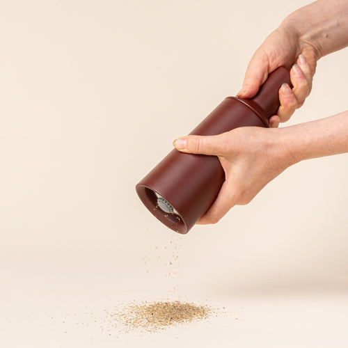 A hand holds out a tall cylindrical sculptural salt and pepper grinder in a dark red on a light background, milling salt