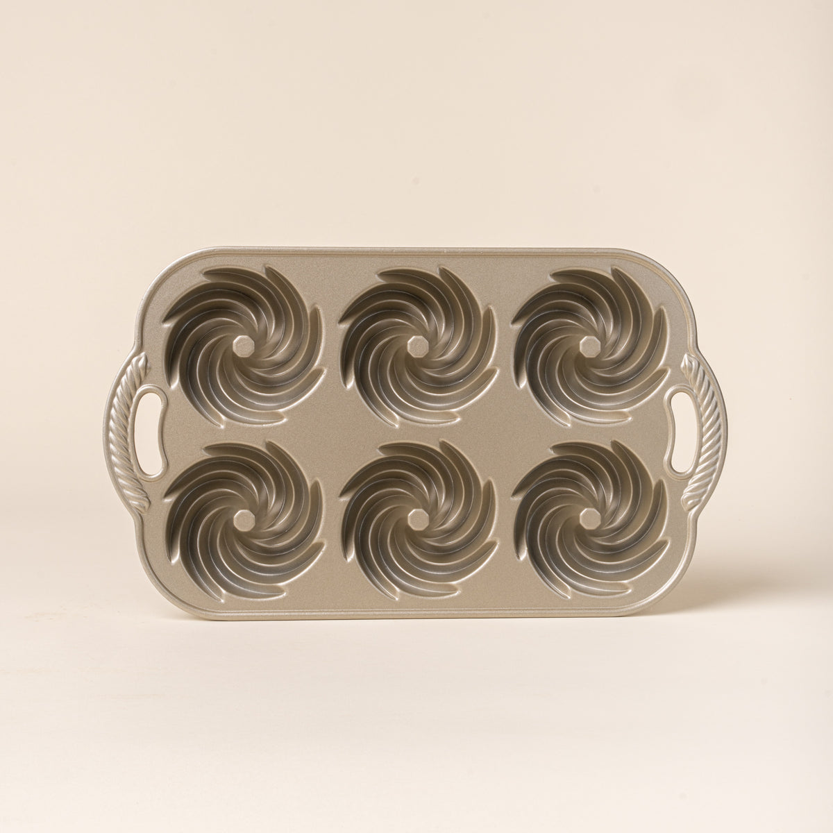 Top view of a silver metal baking pan with six spiral-shaped mini Bundt cake molds and decorative handles on both sides.