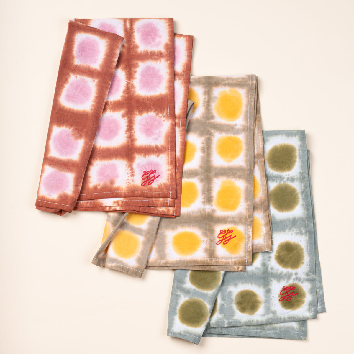 Three folded tie-dye tea towels in pink, yellow, and green colorways with embroidered red details.