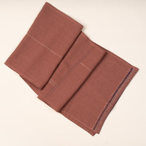 A rust-brown table runner, partially folded to showcase contrasting hand-stitched details in navy and cream.