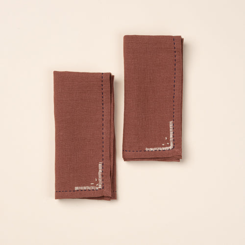 Two folded rust-brown cloth napkins featuring dark blue running-stitch borders and small white embroidered corner details.