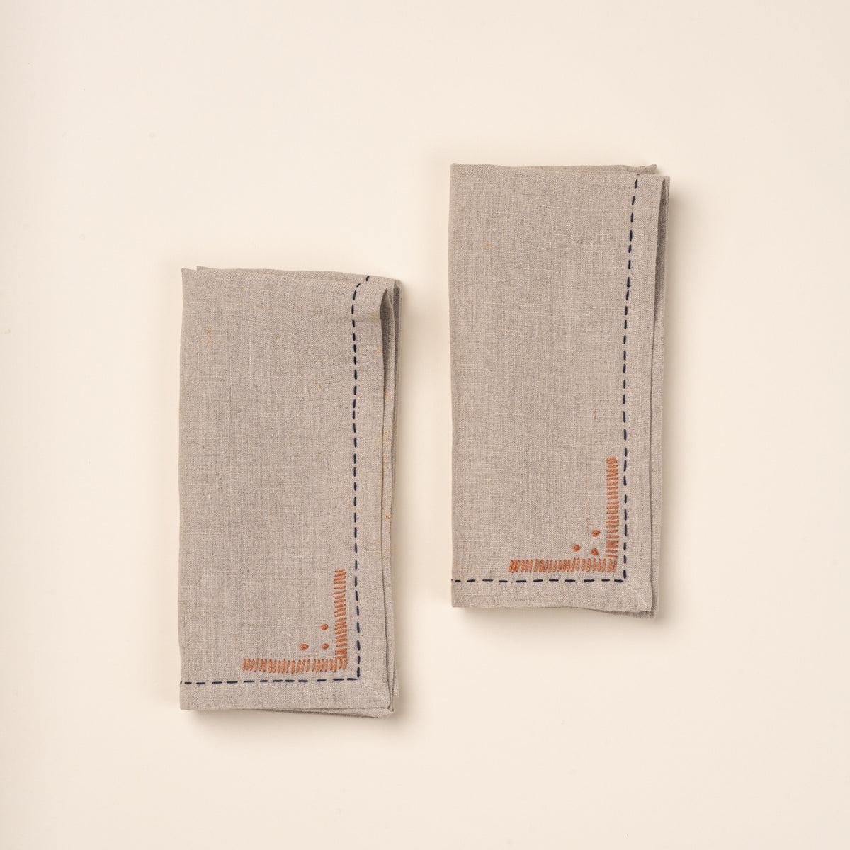 Two folded natural linen napkins featuring dark blue running-stitch borders and small rust-orange embroidered corner details.