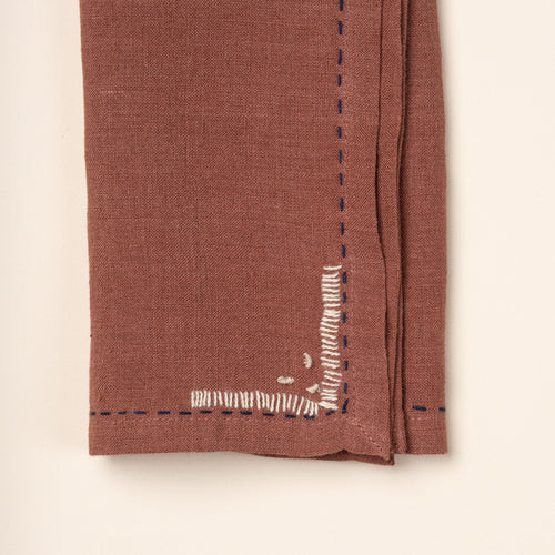 Closeup of a corner of a rust-brown cloth napkins featuring dark blue running-stitch borders and small white embroidered corner details.
