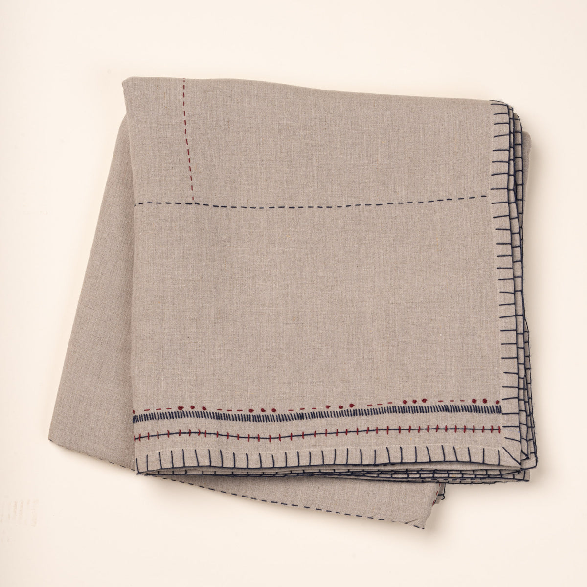 Folded natural linen tablecloth with hand-stitched navy and rust detailing and a decorative geometric border.