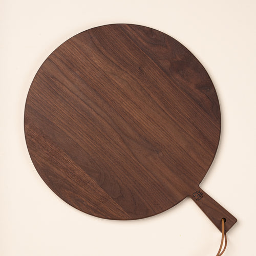 A large round walnut serving board with a short handle and leather hanging loop, shown held against a warm cream background.