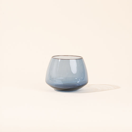 A small dark-blue tinted glass cup with a wide base, on a plain warm off-white background.