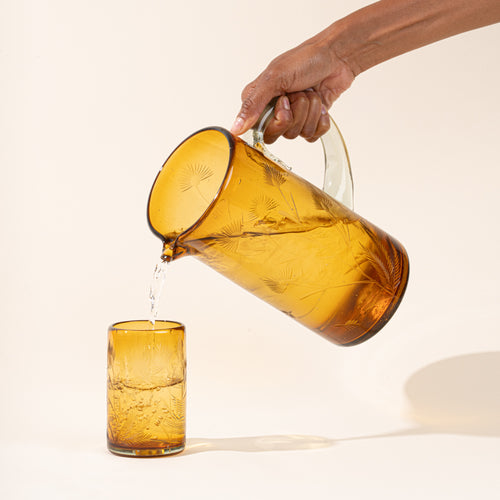 A hand holds a large glass amber pitcher with handle made of recycled glass with etched thistle designs on it pouring water into a matching tumbler.