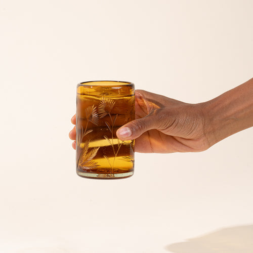 A hand holds an amber tumbler glass made of recycled glass with etched thistle design, filled with water.